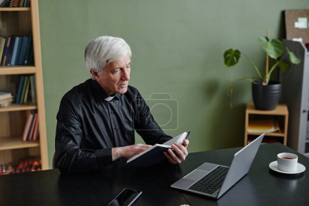 Téléchargez les photos : Portrait de prêtre chevelu blanc lisant la Bible au bureau contre un mur vert, espace de copie - en image libre de droit
