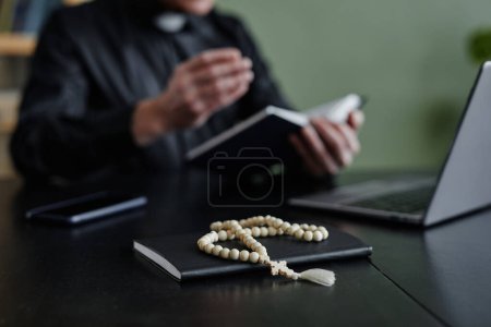 Foto de Primer plano del rosario en el escritorio del lugar de trabajo con un sacerdote irreconocible en segundo plano, espacio para copiar - Imagen libre de derechos