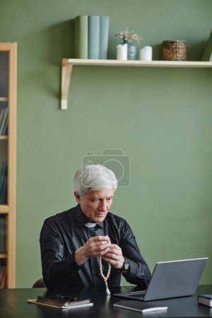 Téléchargez les photos : Portrait vertical de prêtre chevelu blanc tenant le chapelet tout en priant au bureau et en regardant l'écran de l'ordinateur portable, l'espace de copie - en image libre de droit