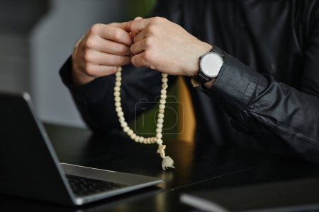 Téléchargez les photos : Closeup of unrecognizable man holding rosary while praying at desk in office, copy space - en image libre de droit