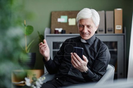 Portrait d'un prêtre aîné souriant utilisant un smartphone et parlant par chat vidéo au bureau, espace de copie