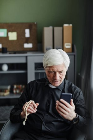 Téléchargez les photos : Portrait vertical d'un prêtre aîné utilisant un smartphone et parlant par chat vidéo au bureau donnant une consultation religieuse en ligne - en image libre de droit
