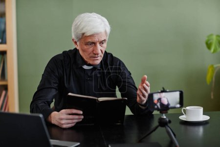 Téléchargez les photos : Portrait d'un prêtre aux cheveux blancs tenant la Bible et enregistrant une vidéo pour un cours religieux en ligne dans son bureau, espace de copie - en image libre de droit