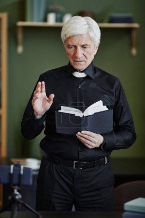 Téléchargez les photos : Portrait vertical d'un prêtre chevelu blanc tenant la Bible et enregistrant une vidéo pour un blog en ligne au bureau - en image libre de droit