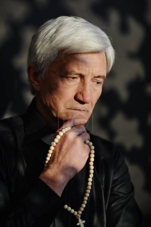 Vertical portrait of white haired senior priest holding rosary deep in thought
