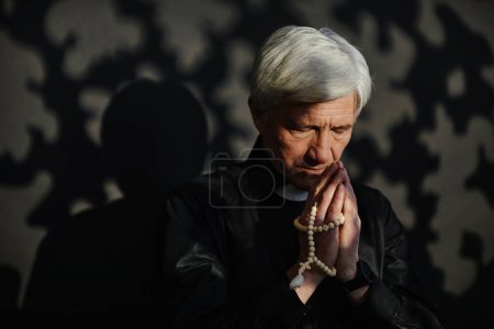 Téléchargez les photos : Portrait dramatique d'un grand prêtre aux cheveux blancs tenant un chapelet et priant dans un éclairage serein, espace de copie - en image libre de droit