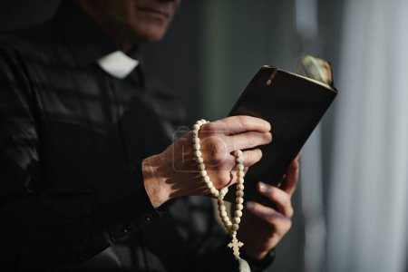 Photo for Side view closeup of unrecognizable priest holding Bible and rosary while praying in dramatic light, copy space - Royalty Free Image