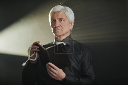 Photo for Dramatic portrait of white haired senior priest holding Bible and praying in dark with god rays, copy space - Royalty Free Image