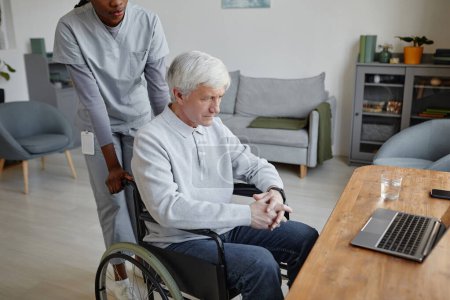 Photo for Side view portrait of white haired senior man in wheelchair at home with nurse assisting - Royalty Free Image