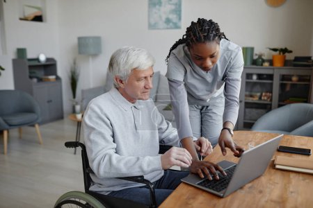 Téléchargez les photos : Portrait d'un aîné handicapé apprenant à utiliser un ordinateur avec l'aide d'une infirmière ou d'un soignant - en image libre de droit