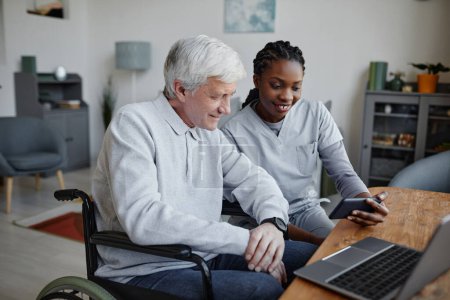 Photo for Portrait of smiling young nurse helping man with disability at home and using smartphone together - Royalty Free Image