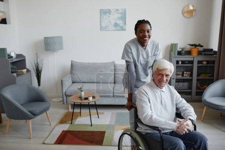 Téléchargez les photos : Portrait d'un homme âgé aux cheveux blancs en fauteuil roulant souriant à la caméra à l'intérieur avec une jeune infirmière aidant, espace de copie - en image libre de droit