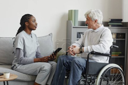 Photo for Side view portrait of black young woman as nurse helping senior in retirement home and using digital tablet - Royalty Free Image
