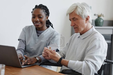 Portrait of white haired senior man holding credit card and learning to use online payment with nurse helping