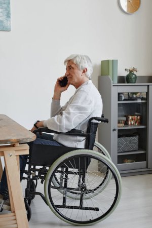 Téléchargez les photos : Portrait vertical latéral de l'homme âgé en fauteuil roulant parlant par smartphone tout en travaillant à la maison - en image libre de droit