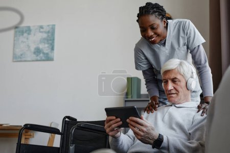 Foto de Retrato de hombre mayor de cabello blanco con discapacidad relajándose en casa y usando tableta con enfermera asistida, espacio para copiar - Imagen libre de derechos