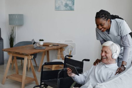 Photo for Portrait of smiling senior man with disability having fun at home and using tablet with nurse assisting, copy space - Royalty Free Image