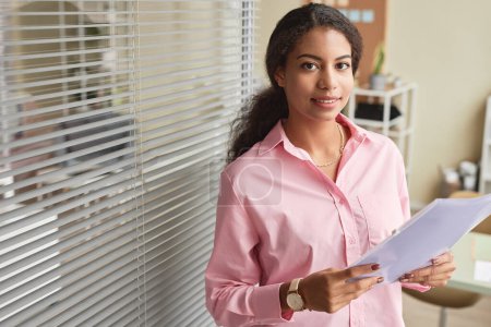 Foto de Retrato de cintura hacia arriba de una joven mujer de negocios negra vestida de rosa en la oficina y sosteniendo el documento sonriendo a la cámara, espacio para copiar - Imagen libre de derechos