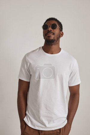 Téléchargez les photos : Portrait minimal d'un bel homme noir portant des lunettes de soleil et posant avec confiance en studio sur fond blanc - en image libre de droit