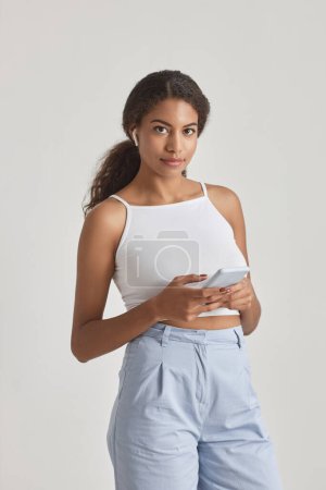 Téléchargez les photos : Portrait franc de la belle afro-américaine posant en studio sur fond blanc tenant smartphone - en image libre de droit