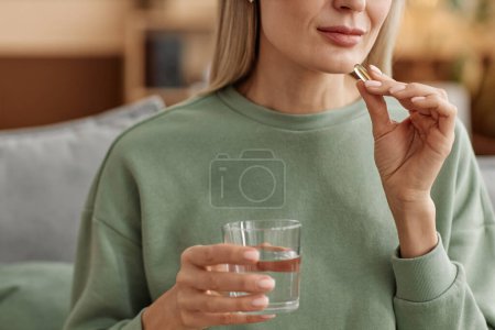 Foto de Primer plano mínimo de la mujer adulta que toma píldora vitamínica y sostiene el vaso de agua en el espacio de copia en casa - Imagen libre de derechos