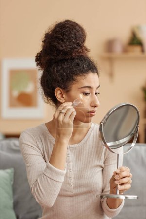 Téléchargez les photos : Portrait vertical latéral de jeune femme appliquant des patchs hydratants et regardant dans un miroir à la maison - en image libre de droit