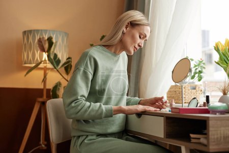Téléchargez les photos : Vue de côté portrait de femme adulte blonde faisant manucure à la table de vanité par espace de copie de fenêtre - en image libre de droit