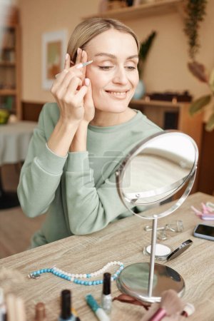 Photo for Vertical portrait of smiling adult woman shaping eyebrows with tweezers looking in mirror at home - Royalty Free Image