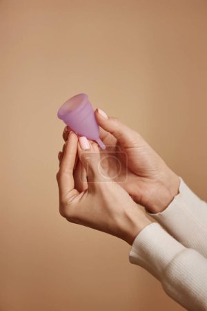 Téléchargez les photos : Gros plan minimal des mains féminines tenant la coupe menstruelle rose sur fond couleur pêche douce - en image libre de droit
