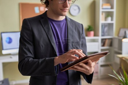 Téléchargez les photos : Gros plan de jeune homme d'affaires utilisant une tablette numérique tout en restant debout à l'intérieur du bureau et en travaillant en ligne, espace de copie - en image libre de droit