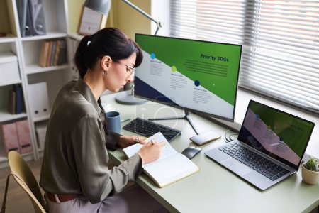 Photo for High angle view at young woman at desk in office or home workplace taking notes in notebook while taking online education course on business strategies - Royalty Free Image