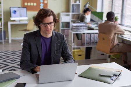 Foto de Retrato de un joven hombre de negocios de pelo largo usando anteojos usando un portátil en el lugar de trabajo de la oficina y sonriendo, espacio para copiar - Imagen libre de derechos