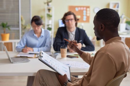 Photo for Side view portrait of black young man holding clipboard while leading business meeting with team in office and discussing project strategy, copy space - Royalty Free Image
