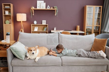 Téléchargez les photos : Portrait de petit garçon utilisant une tablette jouant à des jeux mobiles tout en étant allongé sur un canapé avec chien, espace de copie - en image libre de droit