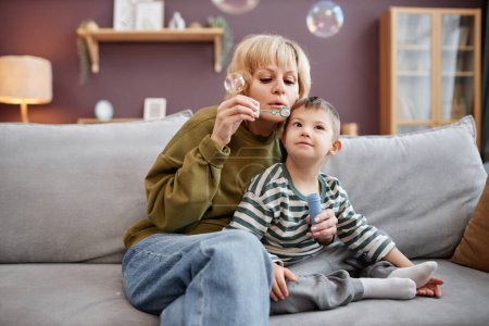 Foto de Retrato de la madre y el niño con síndrome de Down soplando burbujas mientras está sentado en el sofá en casa - Imagen libre de derechos