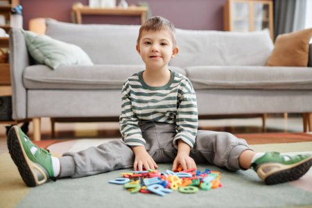 Foto de Retrato de cuerpo entero de niño pequeño con síndrome de Down jugando con juguetes sentados en el suelo y mirando a la cámara - Imagen libre de derechos