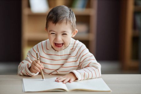 Téléchargez les photos : Portrait d'un petit garçon excité avec un syndrome du duvet dessinant des images assis à table et riant joyeusement, copiant l'espace - en image libre de droit