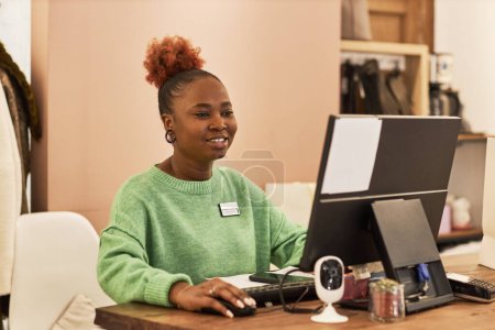 Téléchargez les photos : Portrait de femme noire souriante utilisant l'ordinateur tout en travaillant dans la boutique de vêtements, espace de copie - en image libre de droit