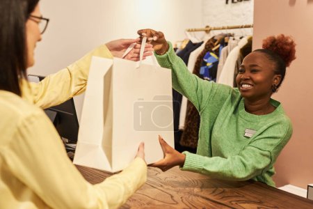 Téléchargez les photos : Portrait de femme noire souriante donnant sac au client en vente libre tout en travaillant dans la boutique de vêtements, espace de copie - en image libre de droit