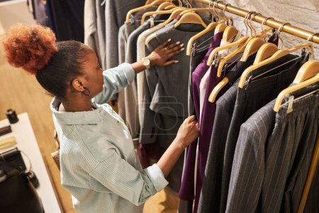Téléchargez les photos : Portrait à grand angle de jeune femme afro-américaine parcourant les vêtements sur étagère dans un magasin de vêtements, espace de copie - en image libre de droit