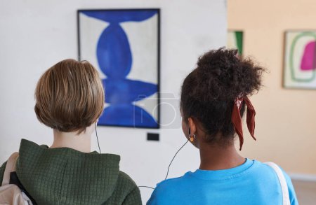 Photo for Back view closeup of two teenagers looking at pictures in modern art gallery or museum sharing earphones with audio guide - Royalty Free Image