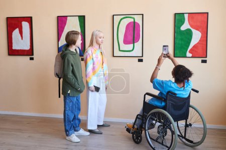 Photo for Back view full length of teenager in wheelchair taking photos of pictures in art gallery or museum while visiting with friends - Royalty Free Image