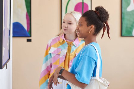 Foto de Colorido retrato de vista lateral de dos chicas adolescentes mirando imágenes en la galería de arte moderno - Imagen libre de derechos