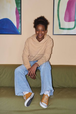 Téléchargez les photos : Portrait vertical de mode d'adolescente noire portant des vêtements décontractés assis sur le canapé, prise de vue avec flash - en image libre de droit