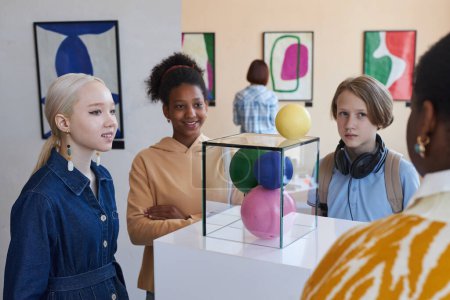Téléchargez les photos : Groupe diversifié d'adolescents souriants écoutant une enseignante ou un guide dans une galerie d'art moderne - en image libre de droit