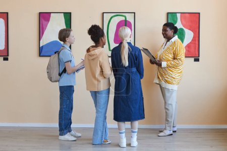 Photo for Colorful back view diverse group of teenagers listening to female teacher in modern art gallery - Royalty Free Image