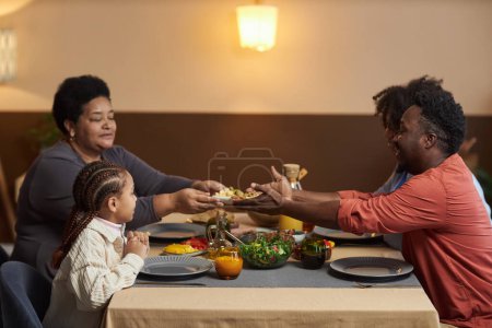 Téléchargez les photos : Portrait latéral d'une famille afro-américaine de trois générations dînant ensemble dans une maison confortable et partageant de la nourriture autour d'une table - en image libre de droit