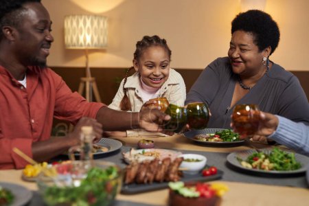 Foto de Retrato de la abuela afroamericana sonriente disfrutando de la cena con la familia en la mesa y tintineo vasos - Imagen libre de derechos