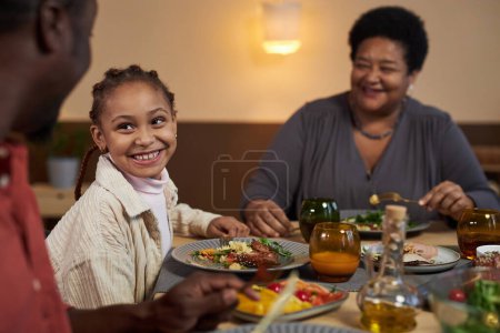 Téléchargez les photos : Portrait de mignonne petite fille noire souriant joyeusement tout en appréciant le dîner avec la famille à la maison espace de copie - en image libre de droit