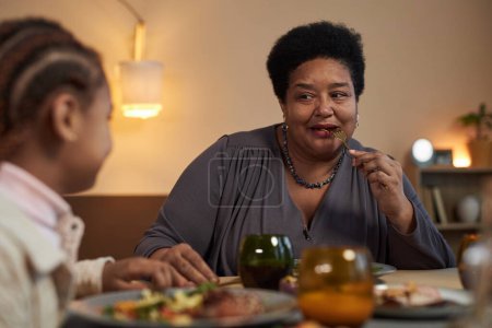 Téléchargez les photos : Portrait d'une femme âgée afro-américaine profitant d'un dîner en famille assise à table et mangeant de la nourriture - en image libre de droit
